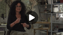La artista Anamaría Devis sentada en su taller o estudio de arte, hablando y haciendo gestos con las manos. Lleva cabello rizado oscuro, gafas de marco claro y ropa oscura. Al fondo se observan estanterías con materiales y piezas artísticas suspendidas. En el centro de la imagen hay un ícono superpuesto de reproducción de video (play).